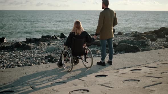 Woman Is Sitting in Wheelchair on Sea Coast and Holding Hand of Her Husband