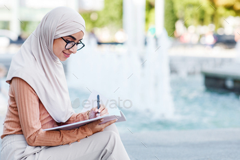 Cheerful cute young middle eastern female in hijab writing in notebook outdoor / Photodune, Misc