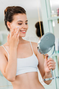 smiling young woman holding mirror and applying facial cream in bathroom / Photodune, Misc