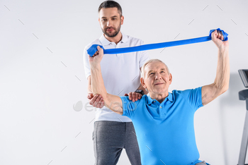 portrait of rehabilitation therapist assisting senior man exercising with rubber tape on grey / Photodune, Misc