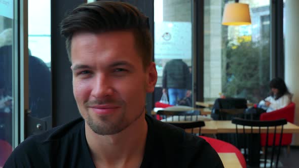 A Young Handsome Man Sits at a Table in a Cafe, Looks Out a Window, Then at the Camera and Smiles