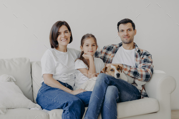 Happy friendly family of mum, dad and daughter pose on comfortable sofa with pet / Photodune, Misc