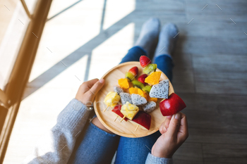 Top view image of a woman eating and holding a wooden plate of fresh mixed fruits on skewers / Photodune, Misc