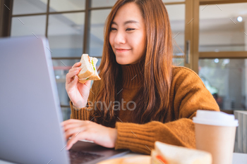 An asian woman holding and eating whole wheat sandwich while working on laptop computer / Photodune, Misc