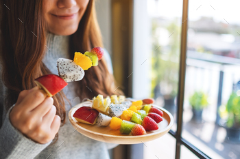 Closeup image of an asian woman holding and eating a fresh mixed fruits on skewers / Photodune, Misc
