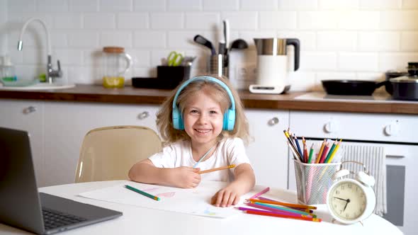 Little Blonde Girl in Headphones and with a Laptop Sitting at a Table in the Kitchen and Smiling