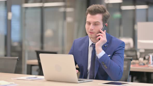 Angry Businessman Talking on Phone While Using Laptop in Office