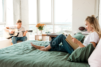 young woman reading book on bed while boyfriend playing acoustic guitar at home / Photodune, Misc