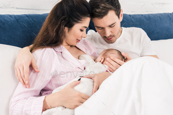 young mother breastfeeding little baby with husband near by on bed at home / Photodune, Misc