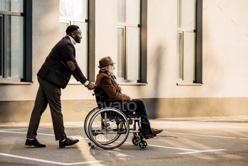 side view of senior disabled man in wheelchair and handsome african american cuidador riding by / Photodune, Misc