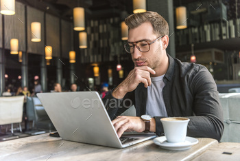 handsome young freelancer working with laptop in cafe with cup of coffee on table / Photodune, Misc
