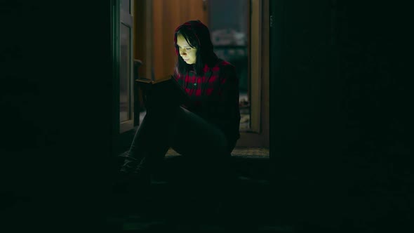 A Beautiful Woman Reads a Book at Night, Sitting on the Porch of a House. Student Leafing Through a