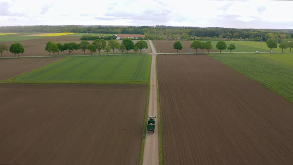 Fly over a tractor which is driving along a argicultural countryside with fields at a rural landscap