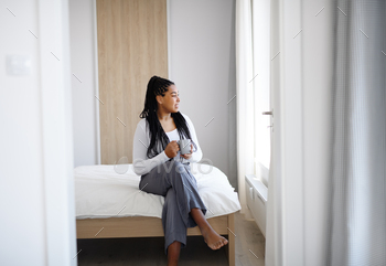 Happy teenage girl with cup of tea sitting indoors in bedroom at home, looking out of window / Photodune, Misc