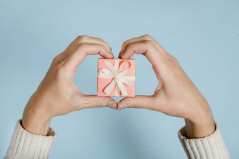 unteer concept with female hands and pink gift box on blue background. Heart shaped female fingers and pink gift box close up.
