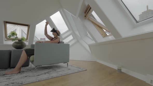 View of Young Blond Woman Sitting on the Side of Arm Chairs Using Tablet Inside of Room in a Cube