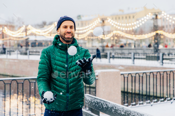 Happy smiling bearded male in warm jacket and hat plays snowballs as stands against beautiful lights / Photodune, Misc