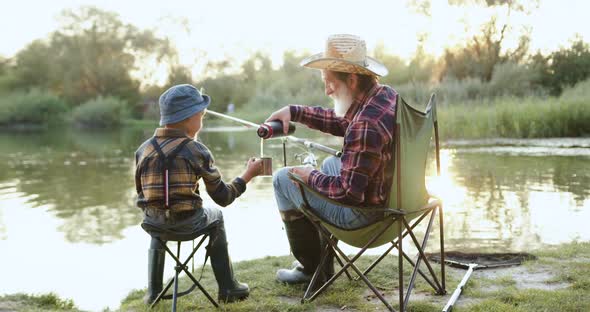 Old Bearded Granddad Filling the Mug with Tea his Handsome Small Grandson while Fishing on the Pond