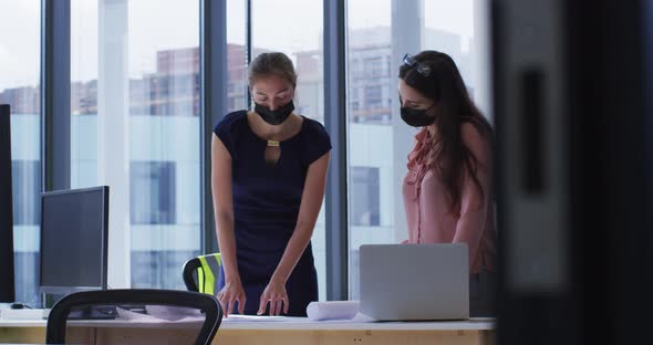 Two diverse female colleague wearing face masks standing at table looking at blueprints and talking