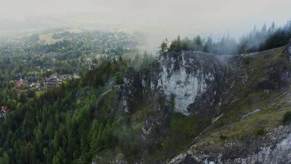 A Village Among a Forest at the Foot of a Sheer Rocky Cliff Covered with Clouds