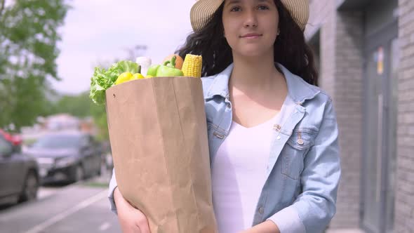 A Young Attractive Woman in a Denim Jacket and Hat Carries a Grocery Bag While Having a Good Mood