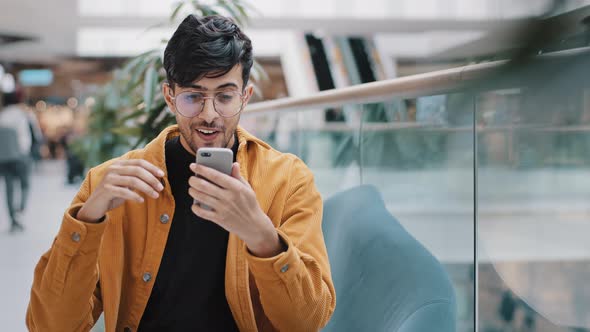 Young Indian Guy Sitting Looking at Smartphone Screen Reading Email Receiving Unexpected Good News