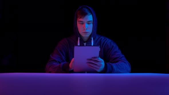 Young Man in a Hood with a Tablet. Hacker Makes a Hack Through the Tablet. Blue and Red Light Falls