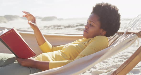 Happy african american woman reading and lying in hammock on sunny beach