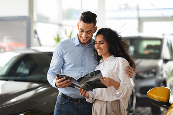 car salon, choosing new family automobile, handsome middle-eastern man and curly woman standing among luxury autos in showroom, holding brochure
