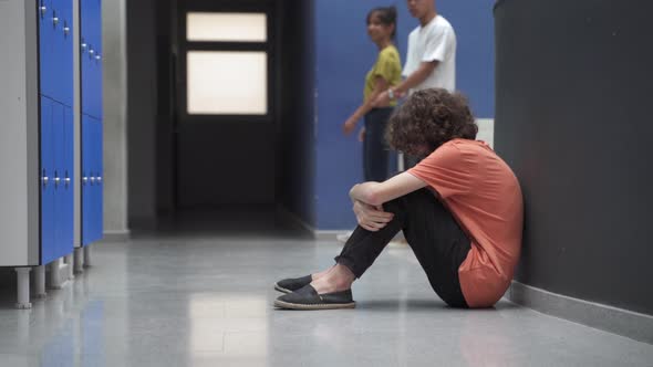 Sad Teenager Sitting Alone on Floor Victim of Bullying While Classmates Ignore Him