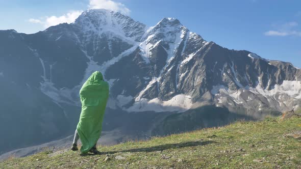 Person in Green Sleeping Bag Jumps Up Against Snowy Mountain