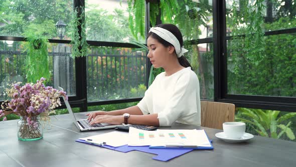 young smiling asian woman working on laptop while enjoying drinking coffee in living room at home.