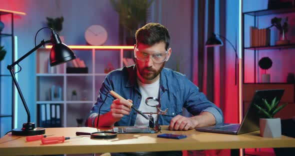 Man in Protective Glasses Soldering Small Parts of Panel Using Soldering Iron and Looking at Camera