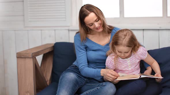 Caring Young Mother and Cute Girl Reading Book Together Sitting on Couch