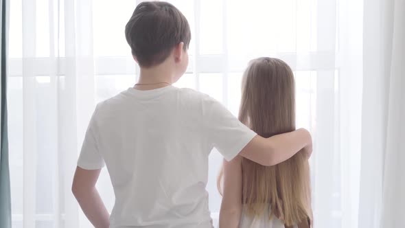 Back View of Caucasian Boy and Girl Standing Together and Looking Out the Window. Brunette Guy in