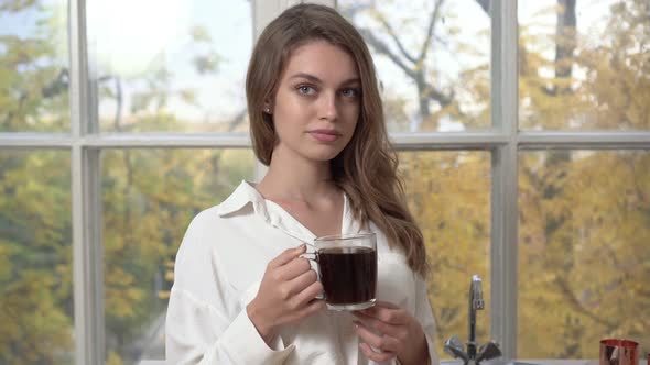 A Young Woman in a White Shirt Drinks Coffee on an Autumn Morning Against the Background of a Window