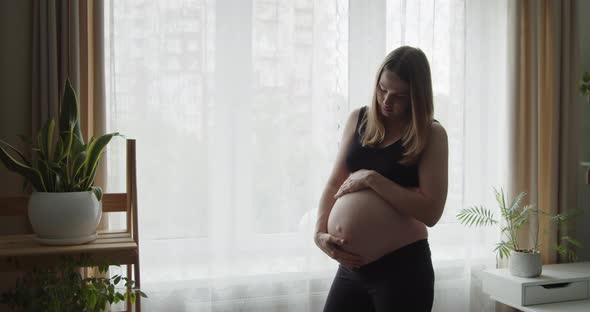 A Young Pregnant Woman Strokes Her Belly with Her Hands Near the Window