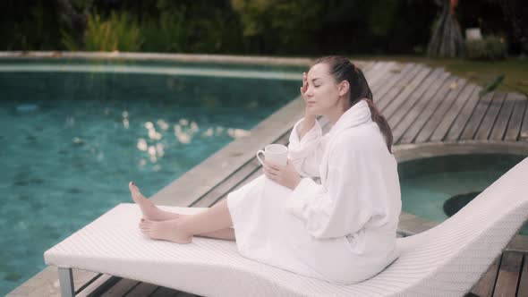 Tranquil Lady in Bathrobe with Mug Sits on Couch Near Pool