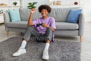 deogame on playstation, having fun at home. Positive Afro teenager with joystick sitting on floor, playing computer arcade. Lockdown hobbies concept