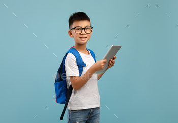  Eyeglasses And Backpack Holding Digital Tablet, Korean Male Child Using Modern Technologies For Studying Online, Standing On Blue Studio Background