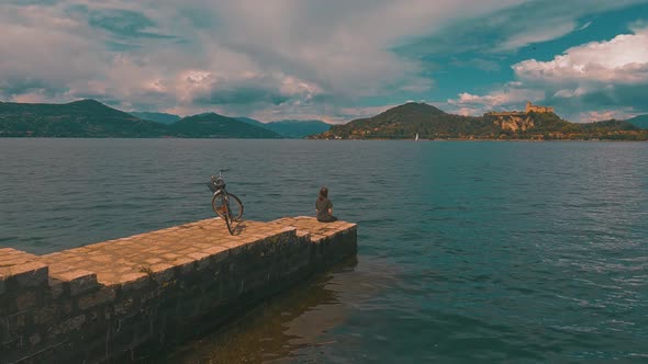 Back view of  lonely woman with bicycle behind sitting on pier edge on Maggiore lake, Italy. Static