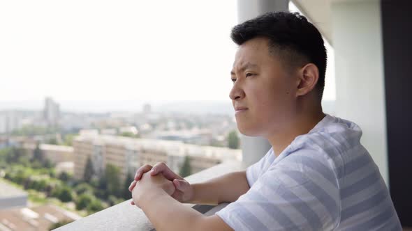 A Young Asian Man Looks Thoughtfully Around on an Apartment Balcony in an Urban Area  Closeup