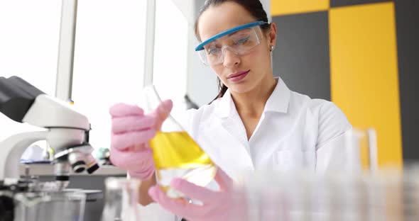 Woman Chemist Looking at Flask with Yellow Liquid in Front of Microscope in Laboratory Movie Slow