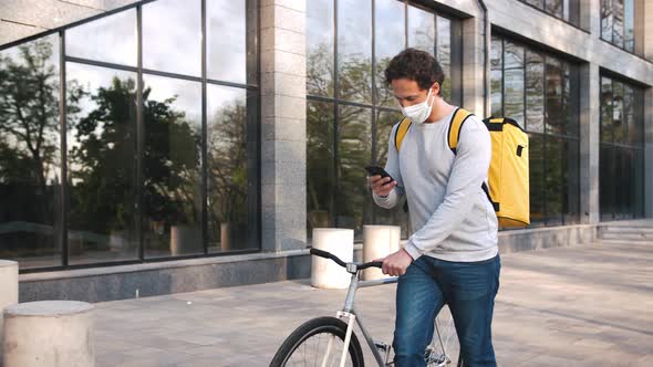 Courier with Yellow Backpack and Protective Mask Walking with Bicycle in City Center and Using