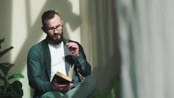 Bearded Young Man Sitting Near Window and Reading a Book at the Sunrise