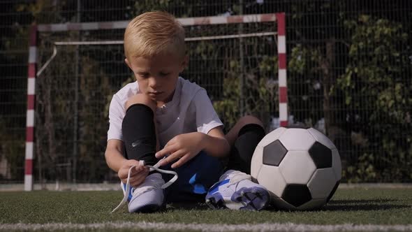 A Little Boy Soccer Player Ties His Shoelaces on a Football Field