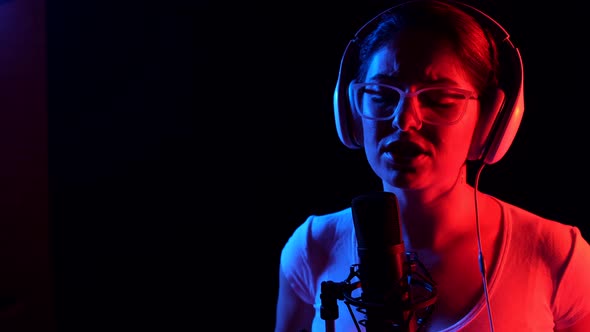 Caucasian Woman in Glasses and Headphones Sings Into a Microphone in Neon Light on a Black