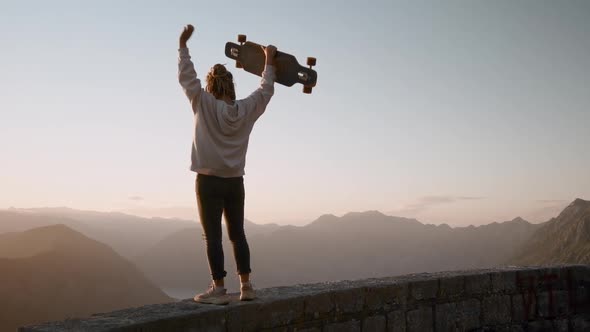 Back silhouette view of Dreadlocks woman longboarder raising hands up feeling sucsess