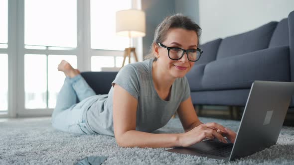 Woman with Glasses is Lying on the Floor and Working on a Laptop