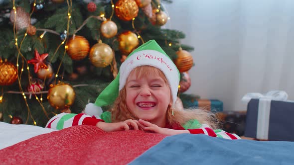 Toddler Girl in Santa Claus Elf Pajama Hiding Behind Bed Playing Hide and Seek Game Christmas Tree